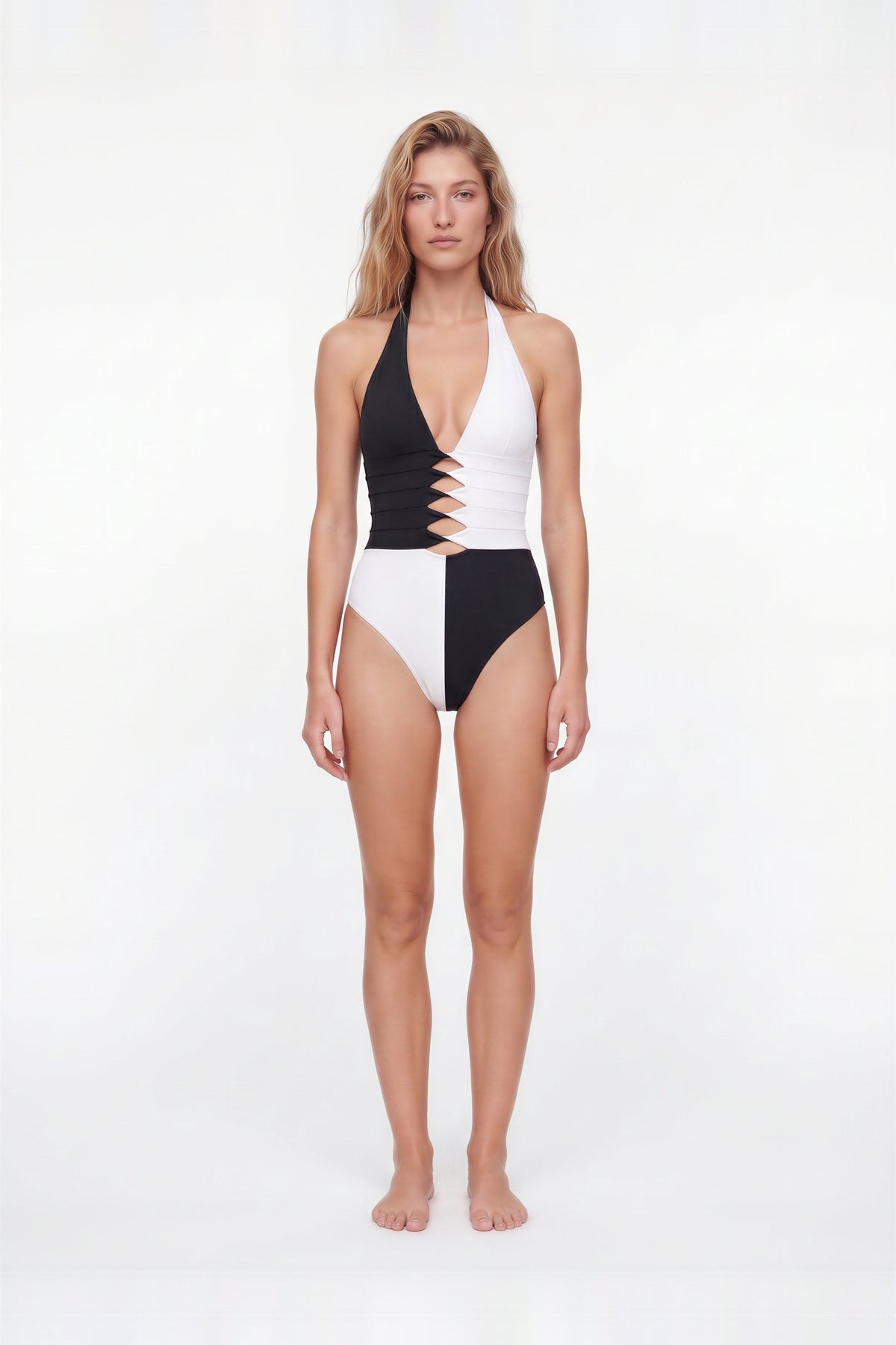 A woman modeling a one-piece swimsuit with a black and white color-blocked design, halter neck, and front cutout details, standing against a plain white studio background.