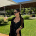 "Model wearing the Itzel Black Kaftan over the Reva Swimsuit, showcasing open-knit design on a beach setting."
