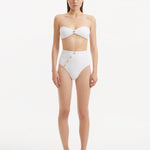 Model showcasing the Baia White Bikini Set from the front, highlighting the strapless bandeau top with gold shell buttons and the minimalist bikini bottom with gold detailing.