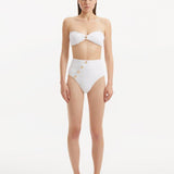 Model showcasing the Baia White Bikini Set from the front, highlighting the strapless bandeau top with gold shell buttons and the minimalist bikini bottom with gold detailing.