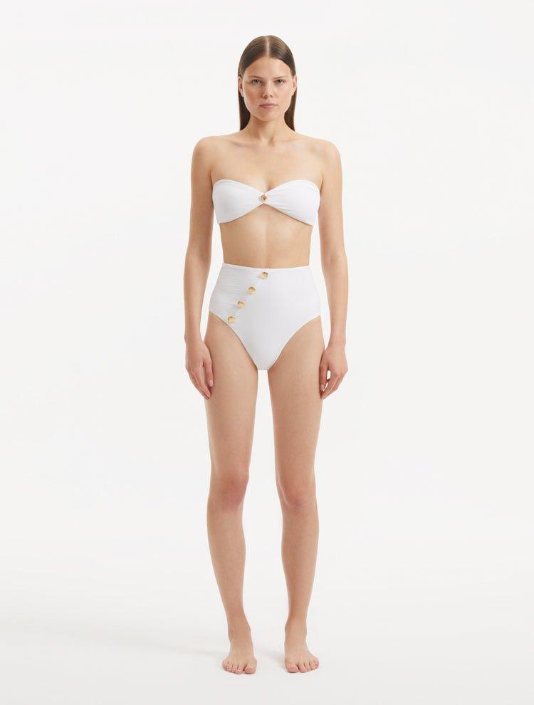 Model showcasing the Baia White Bikini Set from the front, highlighting the strapless bandeau top with gold shell buttons and the minimalist bikini bottom with gold detailing.