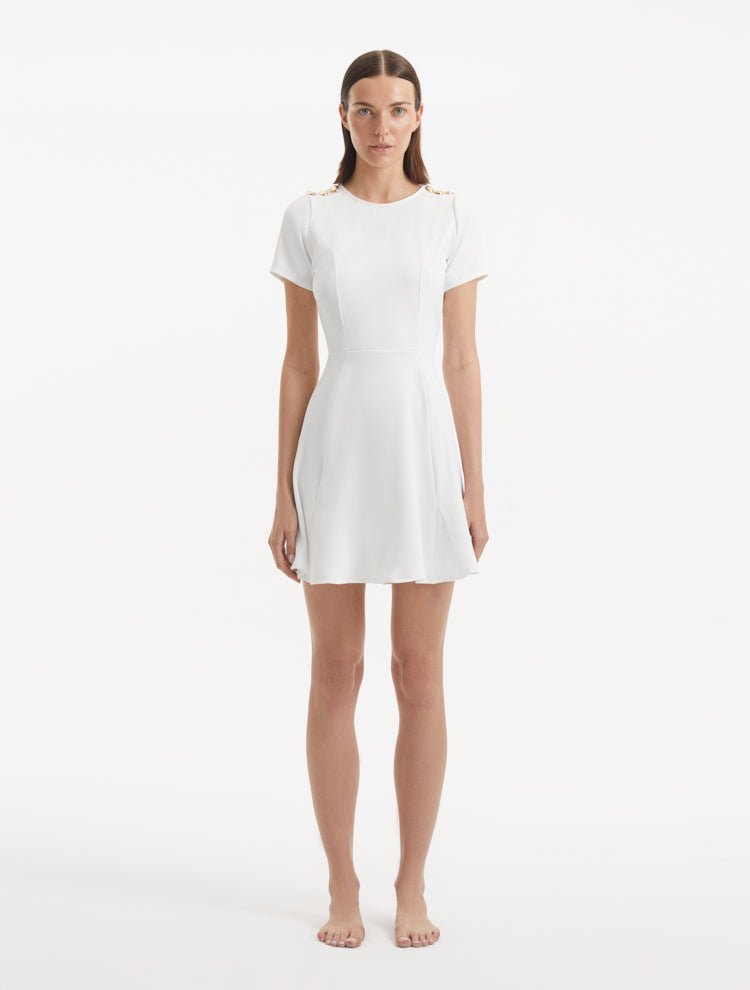 "Model wearing the Derin White Dress from the front, showcasing the knee-length cut, short sleeves, and shell-shaped gold buttons."