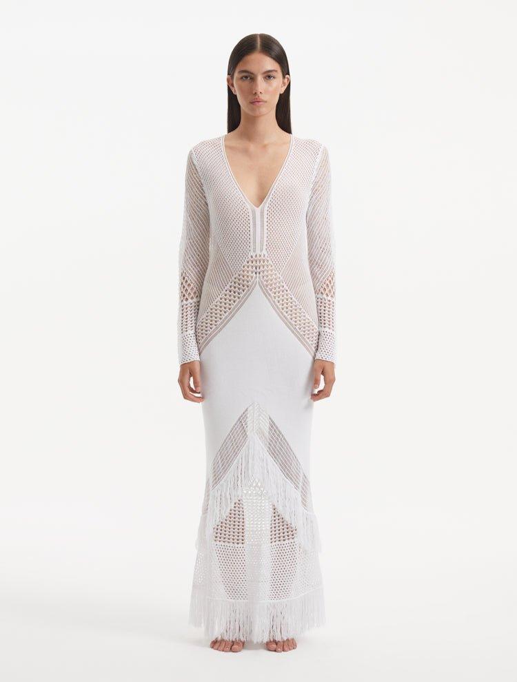 Model showcasing the Elba white dress from the front, highlighting the intricate knitted fabric, V-neckline, and dynamic fringe details. The long semi-sheer sleeves add a sophisticated touch