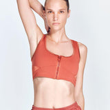 "Fanny Red Ochre Sports Bra front view featuring a square neck, satin matte contrast, and a front zip detail. "