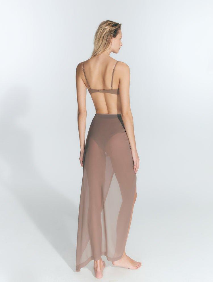 "Back view of a model wearing the Millie Nude Skirt. The skirt features ruched detailing and a front slit with a transparent fabric, offering a sophisticated and chic look. "