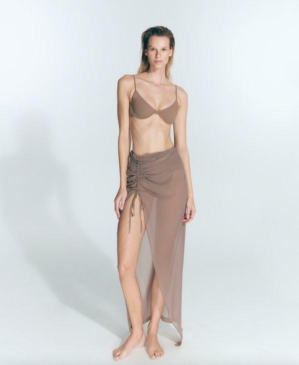 "Front view of a model wearing the Millie Nude Skirt with ruched detailing, a front slit, and transparent fabric.