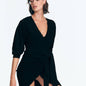 "Front view of the Norma Black Dress on a model, highlighting the V neckline, flowing wrap design, and center slit."